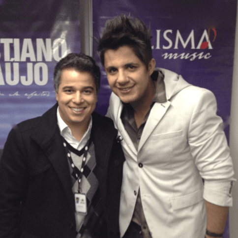 Dois homens sorrindo lado a lado nos bastidores de um show, diante de painéis de divulgação.